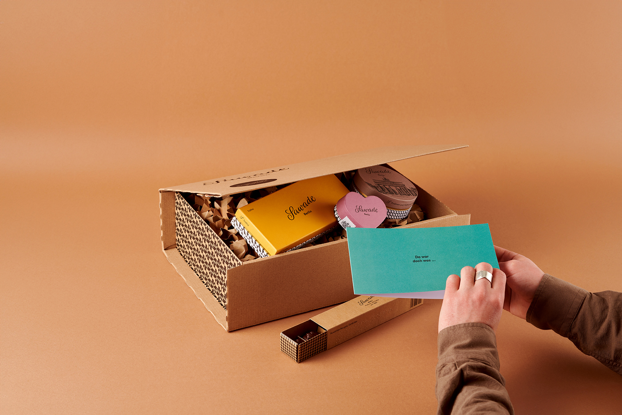 Stimmungsbild: Geöffneter Sawade Versandkarton mit Pralinenschachteln, davor ist eine Hand, die eine Grußkarte hält Stimmungsbild: Geöffneter Sawade Versandkarton mit Pralinenschachteln, davor ist eine Hand, die eine Grußkarte hält