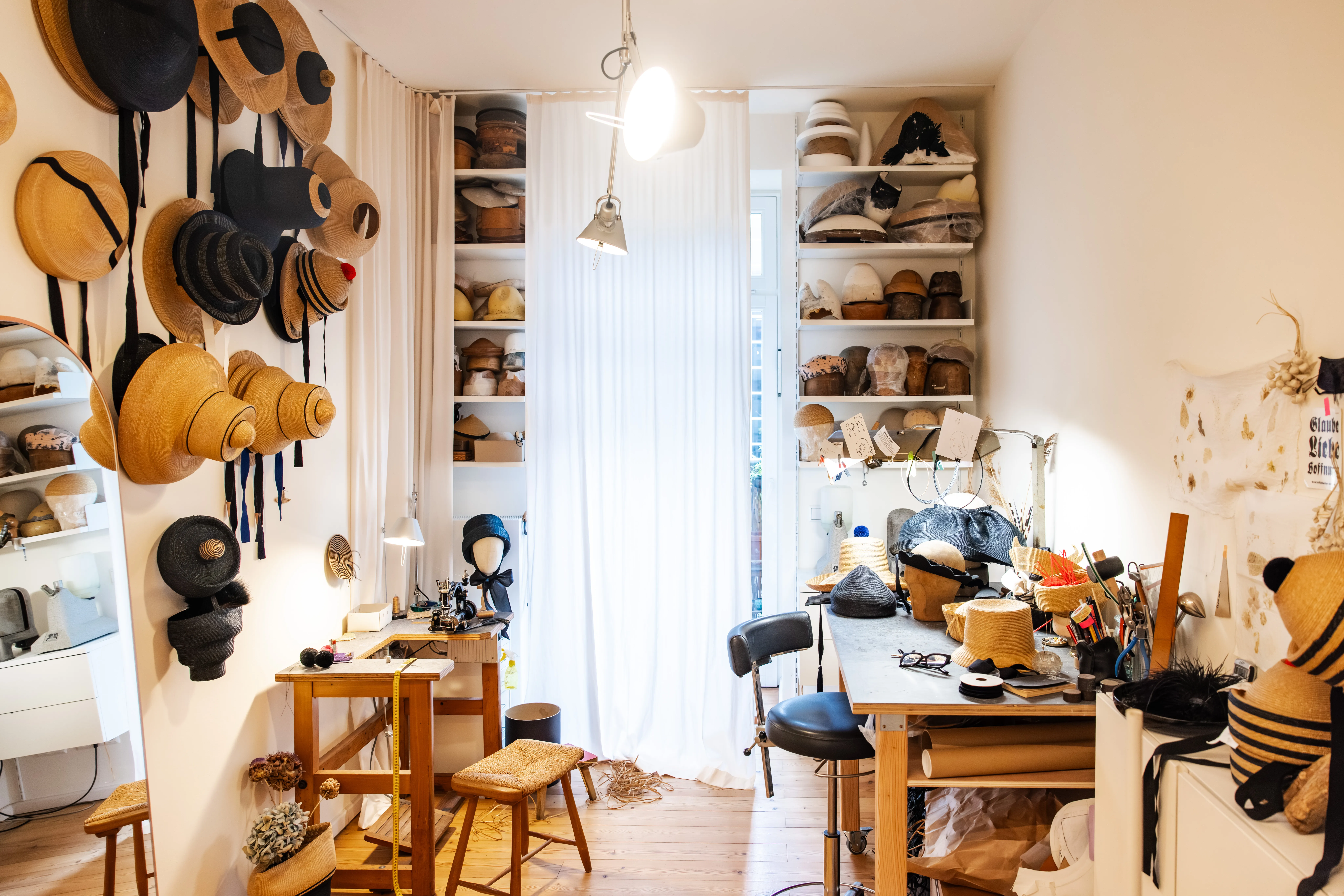 Blick ins Atelier bei SUS Berlin. Hüte an der Wand und in den Regalen. Schreibtisch und Nähmaschine mit Arbeitsutensilien.