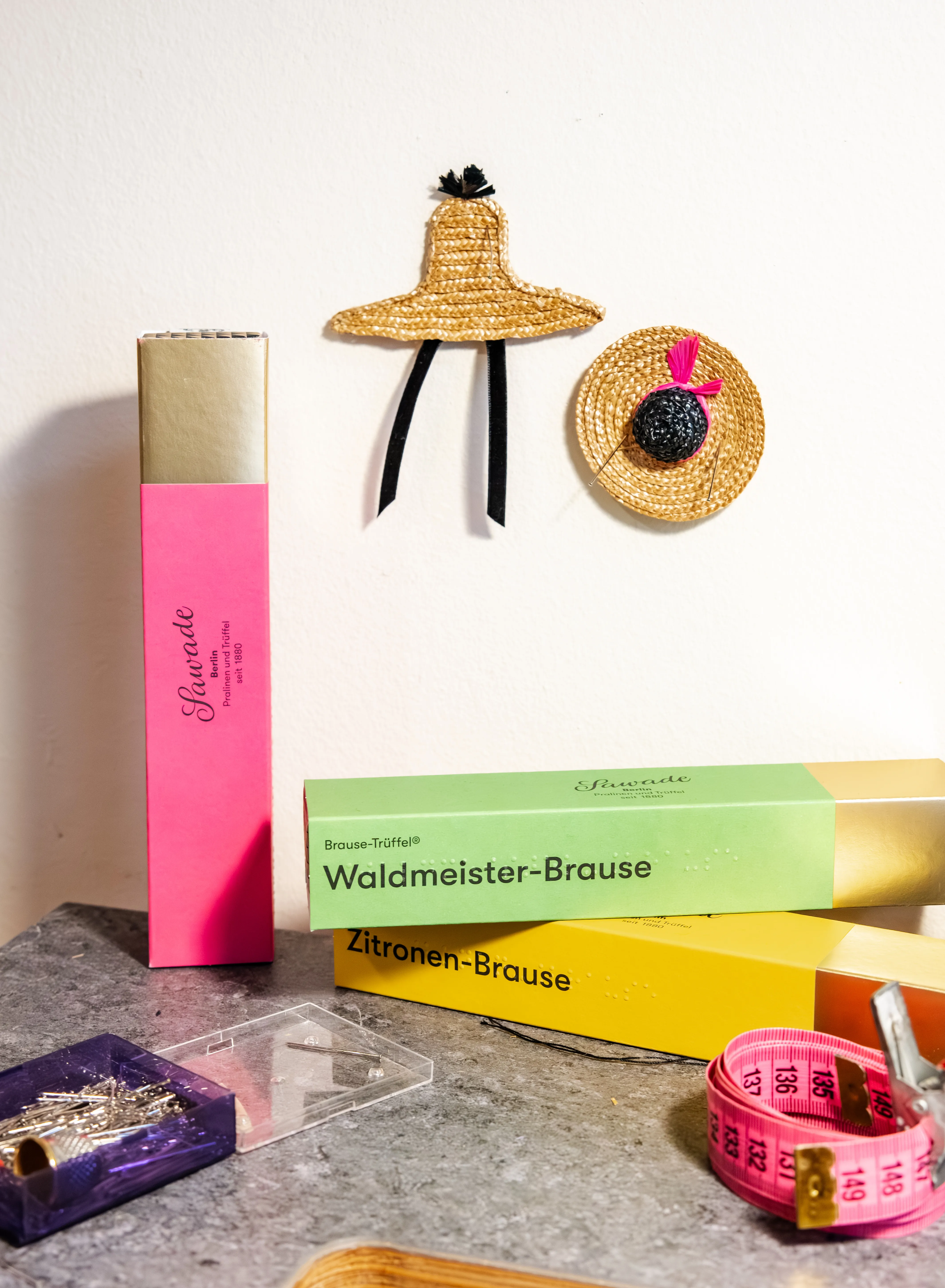 Zu sehen sind drei Stangen Brause-Trüffel in den Farben gelb, gründ und pink sowie zwei kleine Strohhutbroschen, die an der Wand befestigt sind.